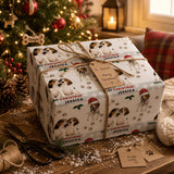 A Christmas gift wrapped in a patterned paper featuring images of beagle dogs, with a note attached that reads "Merry Christmas Jessica". The gift is placed on a wooden surface, with a Christmas tree and other holiday decorations visible in the background.