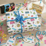 a birthday celebration with a gift wrapped in a white paper with colorful illustrations of a camera, a slice of cake, and other party decorations. The gift is accompanied by a blue ribbon and tags with the name "Adam" written on them.