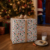 A large gift wrapped in a festive pattern sits on a wooden floor, with a plate of cookies and a glass of milk nearby. In the background, a Christmas tree and a person dressed as Santa Claus can be seen.