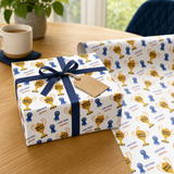 a gift box with a blue ribbon and a tag, placed on a wooden table. The gift box has a pattern of gold trophies and blue ribbons, and the tag reads "Love from James". There is also a roll of wrapping paper with a similar pattern of gold trophies and blue ribbons, and the tag reads "Love from James".