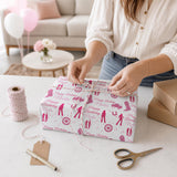 A woman is wrapping a gift with pink and white wrapping paper, surrounded by various crafting supplies and balloons in a room.