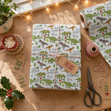 a wrapped gift box with a dinosaur theme, surrounded by holiday decorations and items on a wooden surface.