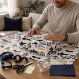 A man is wrapping a gift box with a patterned wrapping paper that has the name "Robert" printed on it. The wrapping paper has a design of military helicopters and other military symbols. The man is using scissors to cut the wrapping paper and a ribbon to secure it. There are also some envelopes and a clock visible in the background.