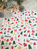 a festive Christmas-themed wrapping paper with various holiday-related icons and decorations, such as pine cones, Christmas trees, ornaments, and a Santa hat. The paper has a pattern of red, green, and white colors, and the text "To: Amelia" is visible, suggesting it is a personalized gift for someone named Amelia.