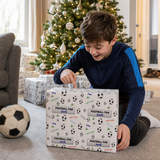 A young boy is sitting on the floor and opening a gift box with a soccer theme, surrounded by a Christmas tree and other holiday decorations.