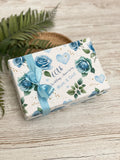 A gift box with a blue ribbon and a floral design, placed on a wooden surface with a plant in the background.