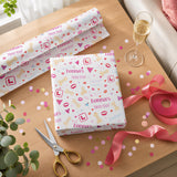 a gift box wrapped in pink and white paper with the word "Donna" printed on it, along with a pair of scissors, a glass of champagne, and confetti on a wooden table.