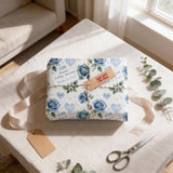 A gift wrapped in blue and white floral paper with a "60th Wedding Anniversary" tag, placed on a white tablecloth with a pair of scissors and some greenery nearby.