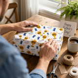 A person is wrapping a gift box with a pattern of gold trophies and ribbons, surrounded by other items such as a coffee cup, a card, and a pair of scissors.