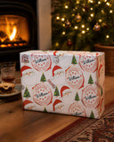 A wrapped gift box with a Christmas-themed design sits on a wooden surface, with a fireplace and a Christmas tree visible in the background.
