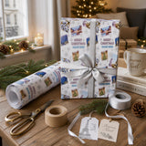 a festive Christmas-themed gift wrapped in a white paper with snowflakes and Christmas trees, along with other holiday decorations such as pine cones and a mug. The scene includes a pair of scissors, tape, and tags, suggesting the preparation of a gift.