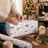 A person is wrapping a gift with a festive Christmas-themed wrapping paper, which has the words "Merry Christmas Alena" printed on it. The person is using scissors to cut the wrapping paper, and there are additional wrapping paper and a tape dispenser visible on the table.