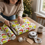 A woman is wrapping a Christmas gift with a patterned wrapping paper featuring tennis balls and a Santa hat. She is using scissors to cut the wrapping paper, and there are other items on the table, such as a cup of coffee and a tape dispenser.