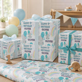 a birthday celebration with a focus on a child's first birthday. There are three wrapped gifts with the name "Olivier" printed on them, along with a wooden toy elephant. The gifts are placed on a table, and there are balloons in the background, creating a festive atmosphere.
