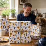 A man is sitting at a table, smiling and looking at a dog, surrounded by wrapped gifts with ribbons and a trophy design.