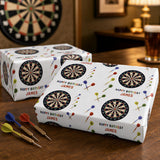 a birthday gift box with a personalized design, including a dartboard pattern and the name "James" printed on it. The gift box is placed on a wooden surface, and there are several darts and a glass of beer visible in the background.
