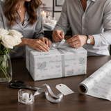 A man and a woman are wrapping a gift together, with the man tying a ribbon around the gift box and the woman holding the gift box.