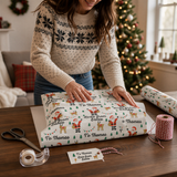A woman in a white sweater is wrapping a Christmas gift with a patterned wrapping paper featuring Santa Claus and reindeer.