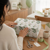 A person is wrapping a gift box with a patterned wrapping paper, surrounded by various items such as scissors, tape, and a mug.