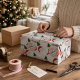 A person is wrapping a Christmas gift with a patterned wrapping paper featuring Santa Claus and other holiday-themed designs. The gift is placed on a wooden table, and the person is using a pair of scissors to cut the wrapping paper. There is also a Christmas tree visible in the background, adding to the festive atmosphere.