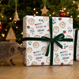 A hedgehog is standing next to a large gift box with a green ribbon, which is decorated with a pattern of hedgehog faces and Christmas-themed elements.