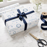 a gift box wrapped in blue and white paper with a ribbon, placed on a marble surface next to a pair of scissors and a vase of white flowers.