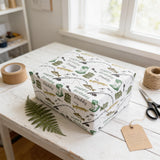 A gift box wrapped in a white paper with a pattern of fishing and fishing-related items, sitting on a white wooden table. There is a pair of scissors and a spool of twine nearby, as well as a tag attached to the gift box.