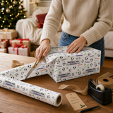 A person is wrapping a gift box with a football-themed wrapping paper, using scissors to cut the paper.
