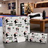 A gift box with a Christmas-themed wrapping paper featuring silhouettes of martial arts fighters and festive text, placed on a wooden table in a room with a Christmas tree and other decorations.