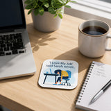 a wooden desk with a laptop, a coffee cup, a notebook, a pen, and a coaster with a cartoon illustration of a person sleeping at a desk.