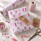 a birthday gift wrapped in pink paper with a cowboy theme, along with other party supplies such as a pair of scissors, a spool of twine, and a tag that says "Hot off the press".