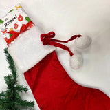 A red Christmas stocking with a white fur trim and a personalized gift tag, along with a small Christmas tree, are displayed on a white surface.
