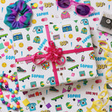 a gift wrapped in a pink ribbon with a pattern of various 90s-themed items, such as sunglasses, a phone, and a skateboard, along with a toy car and a teddy bear.
