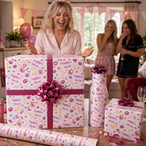 A woman is excitedly opening a large gift wrapped in pink and white paper with the text "Sophie's Hen Do" printed on it, surrounded by other wrapped gifts and party decorations.
