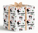 A gift box wrapped in a white paper with a brown bow, featuring a repeating pattern of silhouettes of people engaged in various activities, such as karate fighting and holly leaves. The text "MERRY CHRISTMAS KLEY" is printed on the box, indicating the name of the recipient.