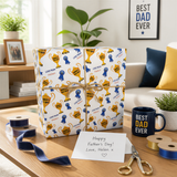 a gift wrapped in a patterned wrapping paper with gold trophies, placed on a wooden table. There is a card with a message for Father's Day, a mug, and some ribbon. The background includes a framed picture and a potted plant.