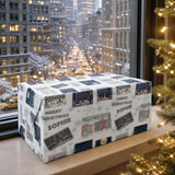 A large, wrapped gift box sits on a windowsill, with a cityscape visible through the window. The gift box is decorated with festive holiday-themed images and text, including phrases like "Merry Christmas" and "Sophie".