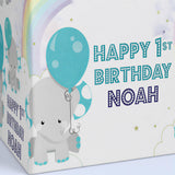 A gray elephant holding a blue balloon, with a rainbow and stars in the background, and the text "HAPPY 1ST BIRTHDAY NOAH" written on the side of the elephant.