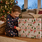 A young boy in a Santa pajama set is excitedly opening a Christmas gift, with a decorated Christmas tree visible in the background.