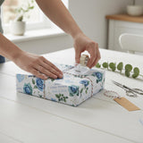 A person's hands are wrapping a gift box with blue and white floral wrapping paper, using a roll of tape to secure the wrapping.