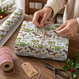 A person's hands are wrapping a gift box with dinosaur-themed wrapping paper, surrounded by holiday decorations such as holly and pine branches.