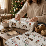 A woman is wrapping a Christmas gift with a patterned wrapping paper featuring images of dogs, surrounded by holiday decorations and a Christmas tree.
