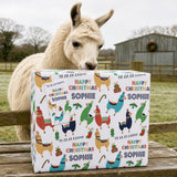 A white llama is standing behind a colorful gift box with a Christmas theme, featuring various llama illustrations and the text "Happy Christmas Sophie".