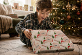 A young boy in pajamas is excitedly opening a Christmas gift wrapped in a patterned paper with Santa Claus designs.