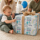 A baby boy is sitting on the floor, playing with a large gift box that has a birthday theme. The gift box is wrapped with a blue and white pattern and has a brown bow on top.