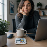 A woman in a gray sweater is sitting at a desk, working on a laptop computer. She has a coffee mug and a coaster with a cartoon character on it. There are also several books and plants in the background, creating a cozy and focused work environment.