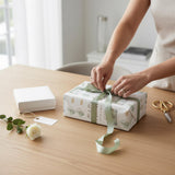 A person is wrapping a gift with a green ribbon on a wooden table, surrounded by a white box, a rose, and a pair of scissors.