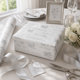 a table setting with a white tablecloth, silverware, and a framed picture. There is a gift box wrapped in white paper with a silver ribbon, and a tag that reads "25 Years".