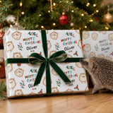 A hedgehog is standing next to a large gift box with a green ribbon, which is wrapped with a Christmas-themed design featuring hedgehog illustrations and the text "Merry Christmas Oliver".