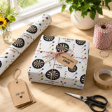 a birthday gift wrapped in a white paper with a dartboard pattern, placed on a wooden surface next to a pair of scissors, a roll of tape, and a vase with flowers.